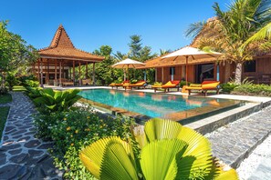 A pool area with lounge chairs and umbrellas, surrounded by tropical plants and a traditional building. - Stunning Joglo in Luxury Style! (Seririt)