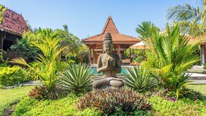 Un jardin paisible, orné d’une statue de Bouddha, d’une végétation luxuriante et d’un bâtiment traditionnel doté d’un toit en tuiles.