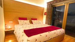Une chambre avec un plafond en bois, un grand lit recouvert d’une literie à motifs, une table de chevet avec une lampe, et une vue sur un paysage enneigé par une porte coulissante.