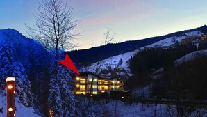 Un paysage enneigé avec un bâtiment illuminé en arrière-plan, un arbre recouvert de neige et un sentier enneigé bordé de lumières.