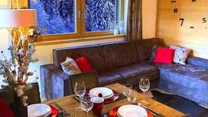 Un coin repas avec une table dressée pour quatre personnes, un plafond en bois, une fenêtre donnant sur des arbres couverts de neige et une horloge murale.