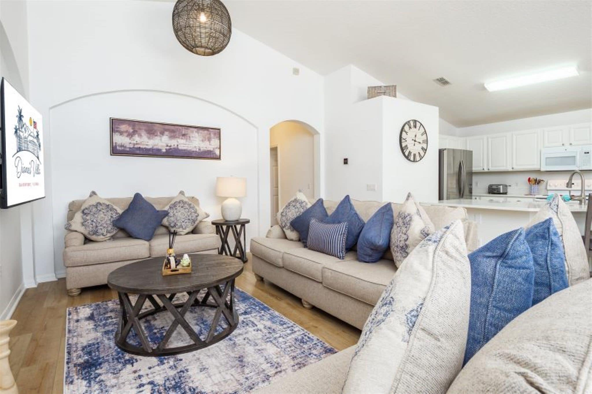A modern living room with a beige sofa, a round coffee table, and a blue patterned rug. There is a kitchen area with white cabinets and a stainless steel refrigerator.