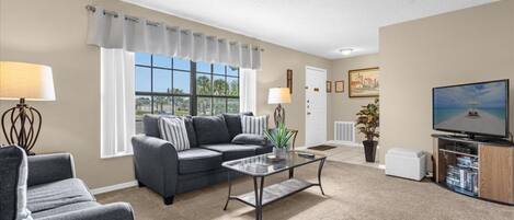 A living room with a grey sectional sofa, a glass coffee table, a flat-screen TV, and a bookshelf.