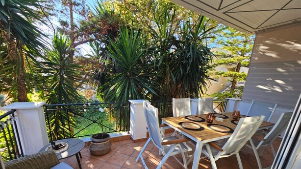 A balcony with a wooden table and white chairs, surrounded by potted plants and a view of trees. - Corfu Dream Holidays Villa Kanoni (Corfu)