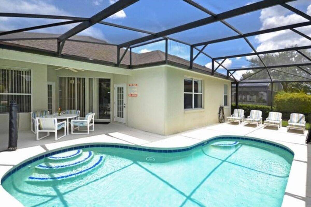 A swimming pool with a glass enclosure, a covered patio with chairs, and a building with a sign.