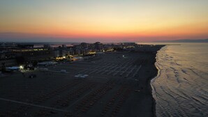 A beach with rows of umbrellas and beach chairs, a promenade with buildings, and the ocean at sunset. - Beautiful Family Apartment in the Center (Margherita di Savoia)