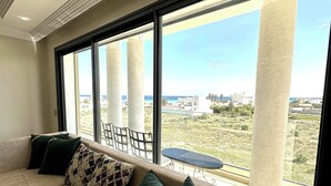 A modern living room with a beige sectional sofa, green and patterned cushions, large windows with a view of a coastal landscape, and a small table.