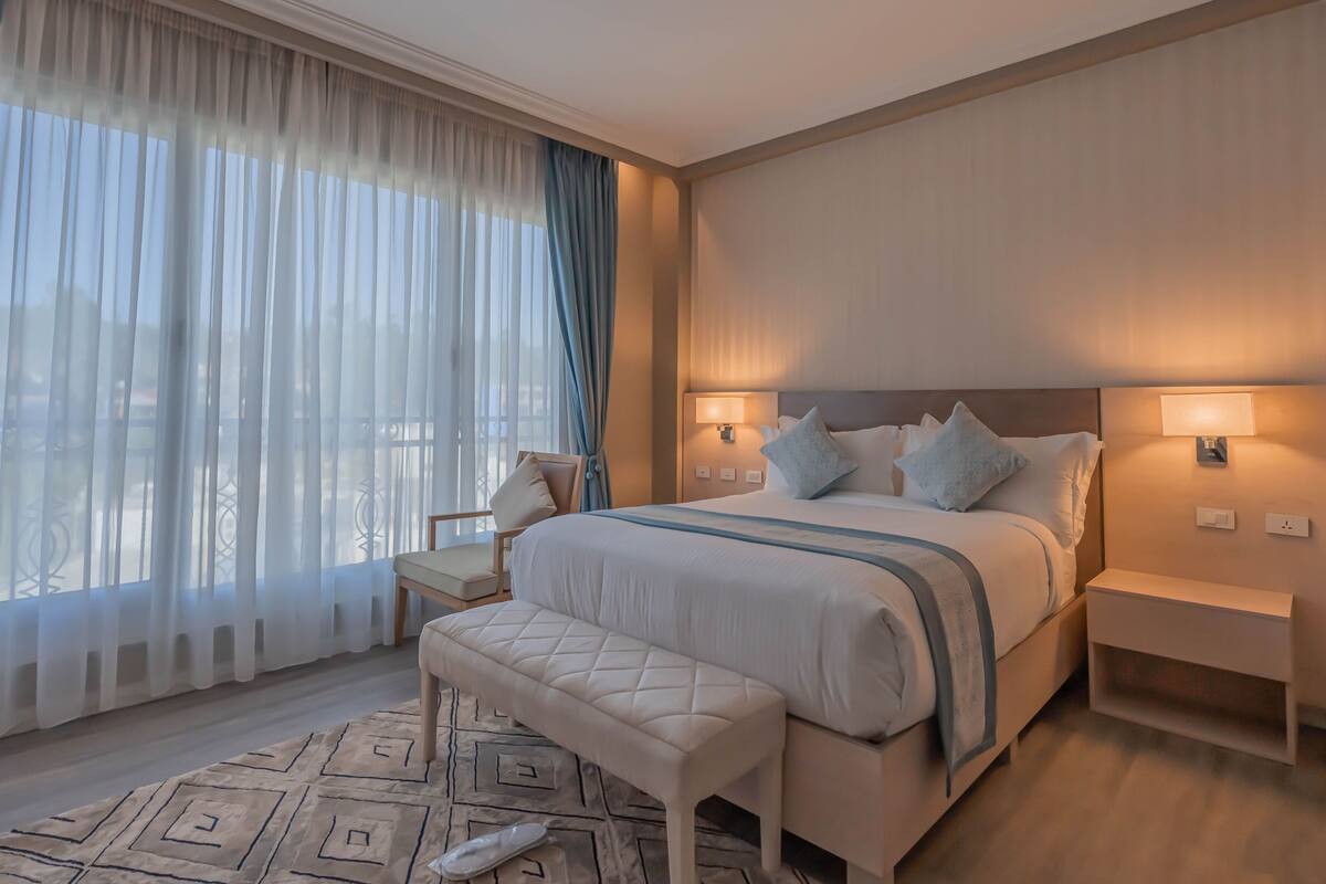 Une chambre d’hôtel avec un grand lit, deux lampes de chevet, un banc et une vue à travers des rideaux transparents.
