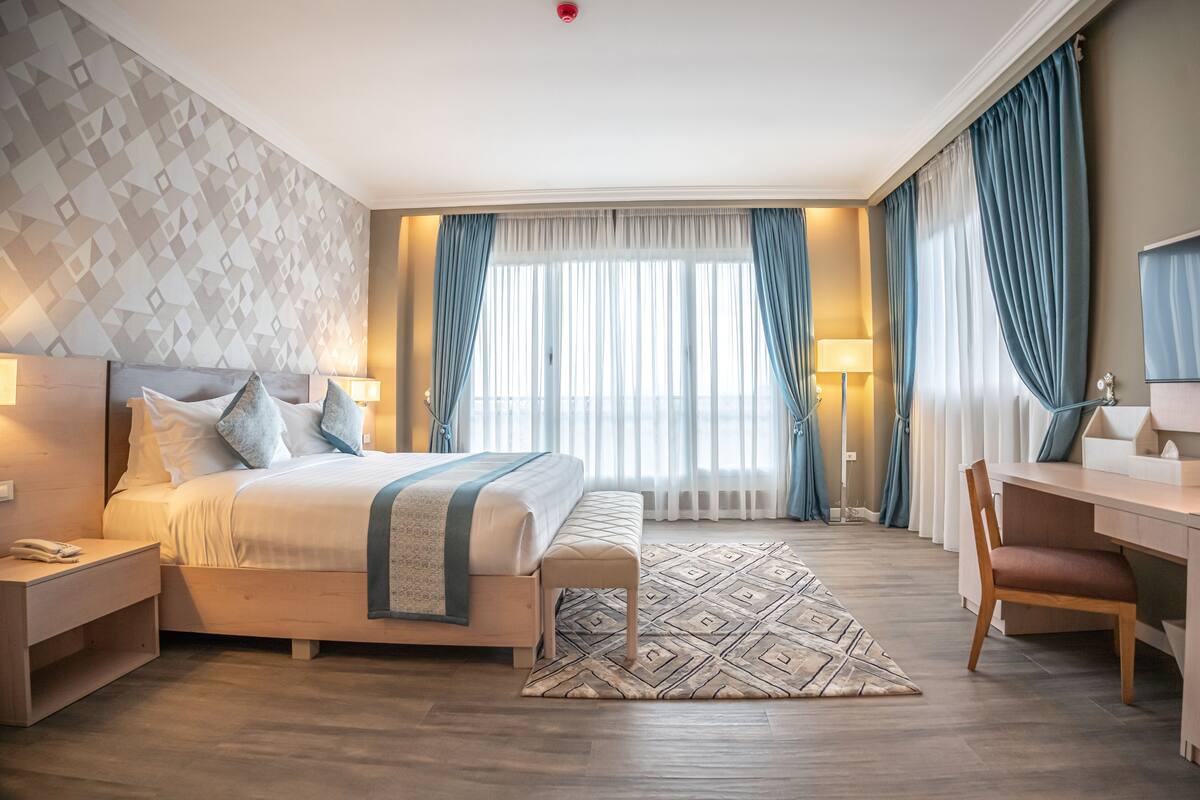 Une chambre d’hôtel comprenant un lit, des tables de chevet, un bureau, une chaise et une grande fenêtre avec des rideaux.