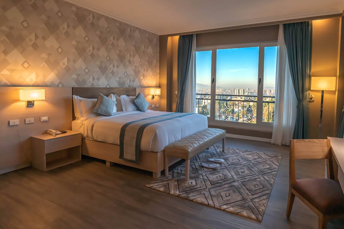 Une chambre d’hôtel avec un grand lit, des lampes de chevet, un banc, une chaise en bois et une vue sur le paysage urbain par une grande fenêtre.