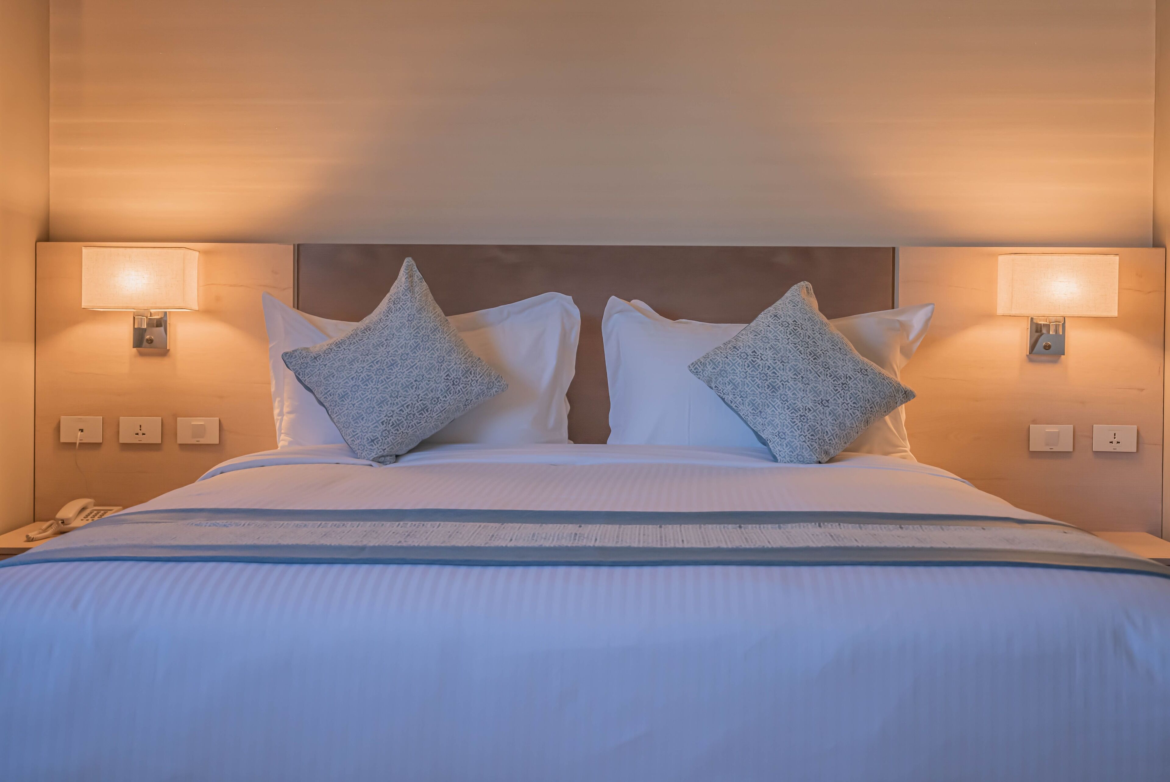A neatly made bed with white bedding and two patterned pillows, flanked by wall-mounted lamps.