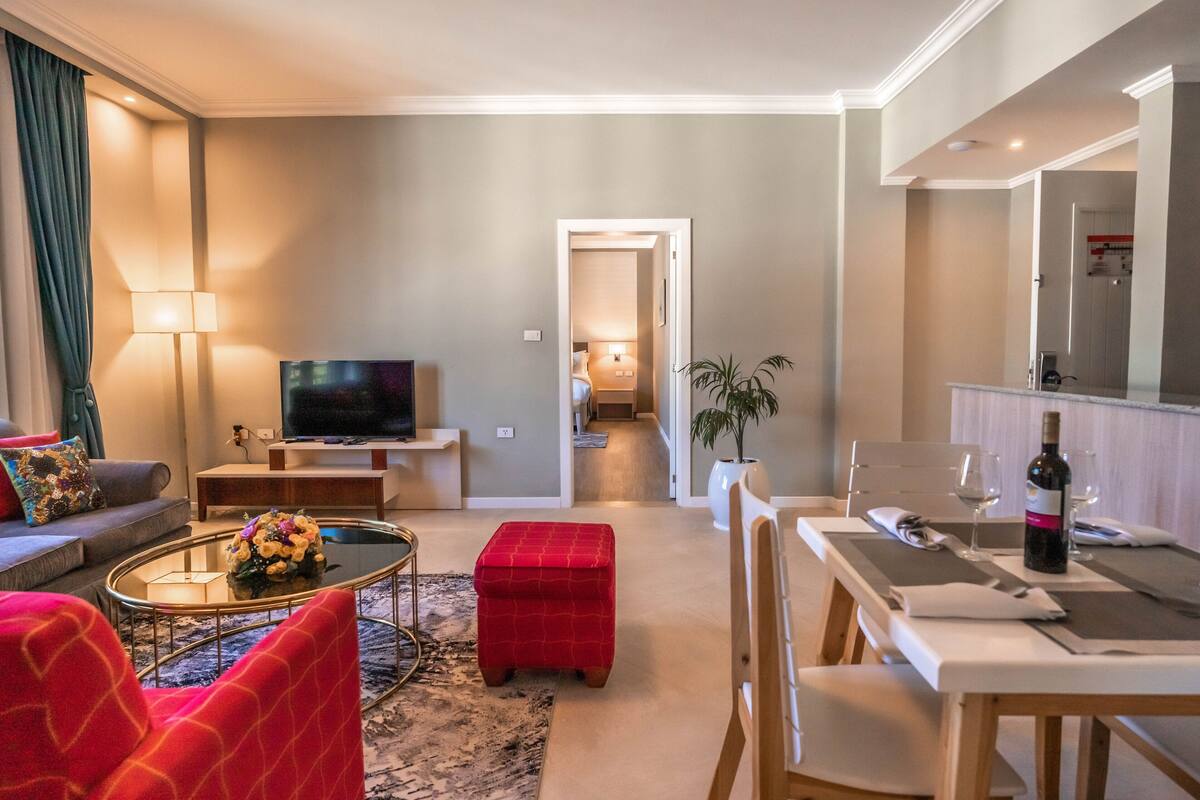 Un salon moderne avec un téléviseur, un pouf rouge et un coin repas avec une table mise pour deux personnes.