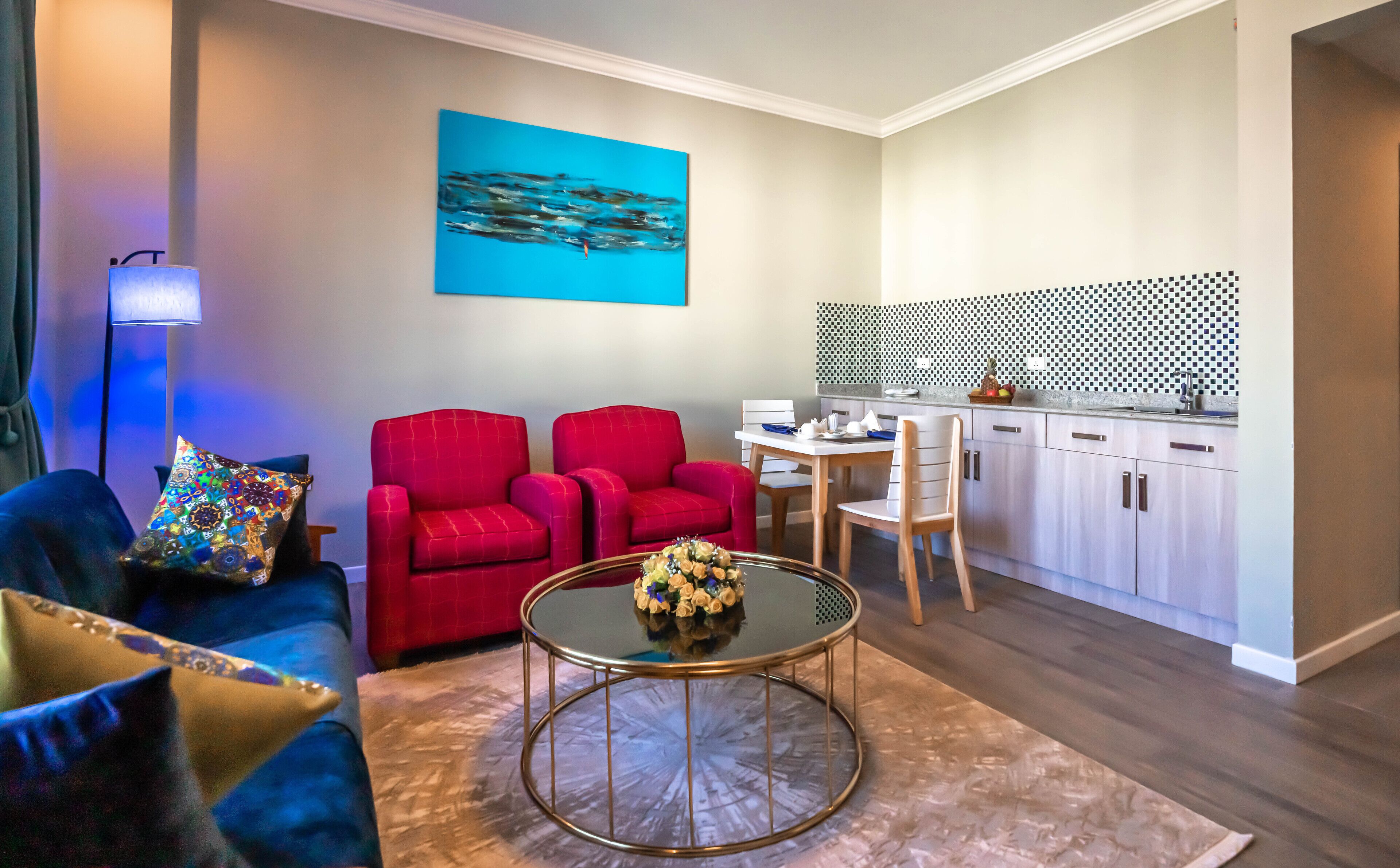 A modern living room with a blue sofa, red armchairs, and a coffee table. A kitchen with white cabinets and a black and white checkered backsplash is visible.