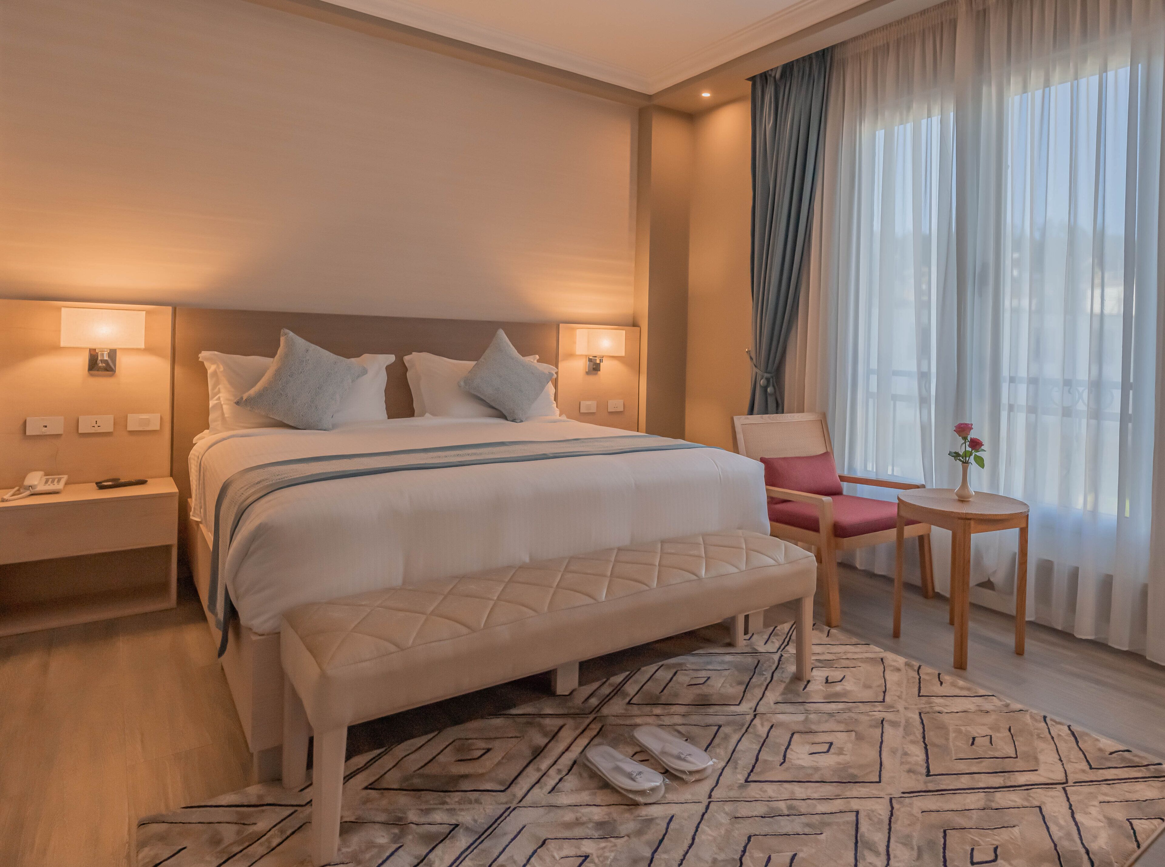 A hotel room with a bed, bedside lamps, a bedside phone, a bench, a chair, a small table with a vase, and a patterned rug.