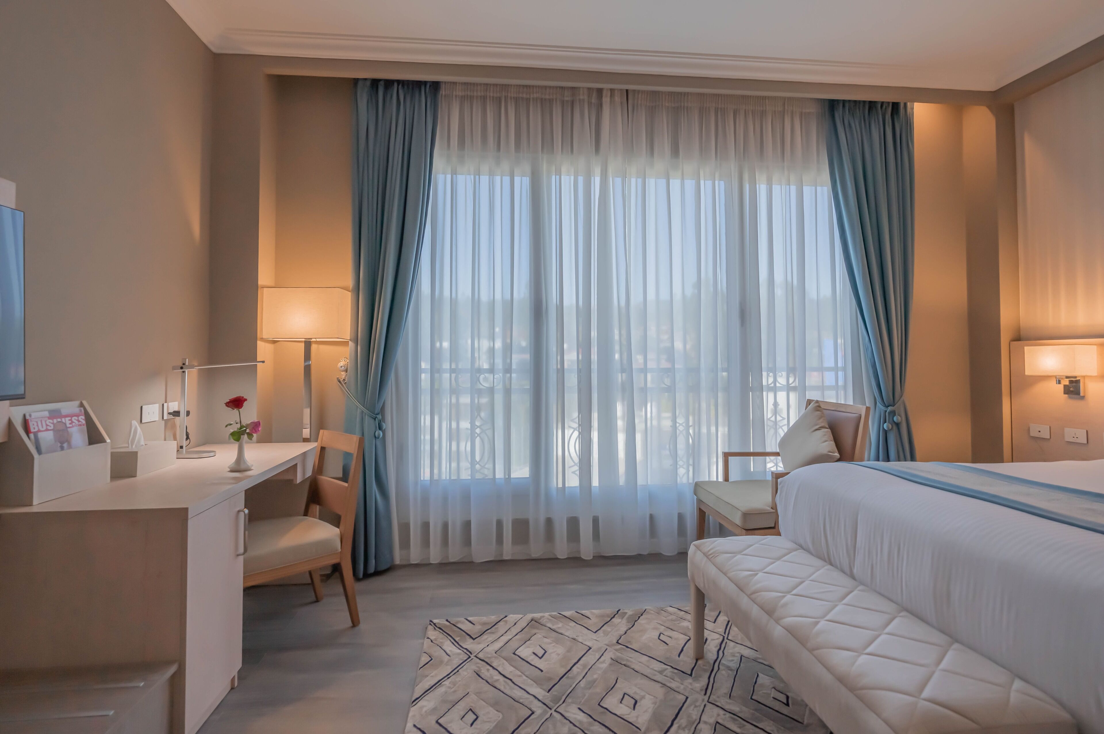 A hotel room with a bed, a desk, a chair, and a window with sheer curtains.