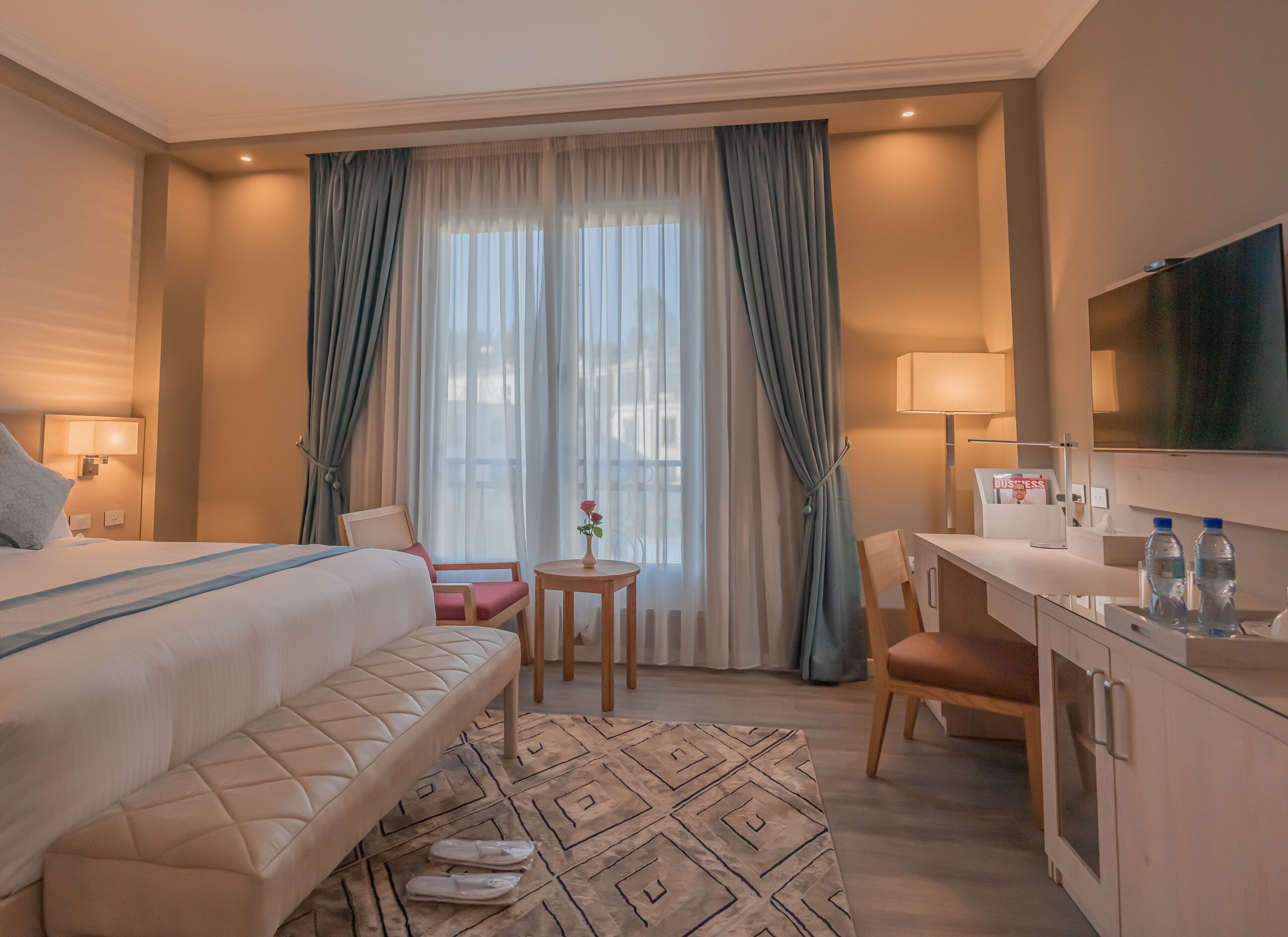 A hotel room with a bed, a desk, a chair, a small table with a vase, and a television.