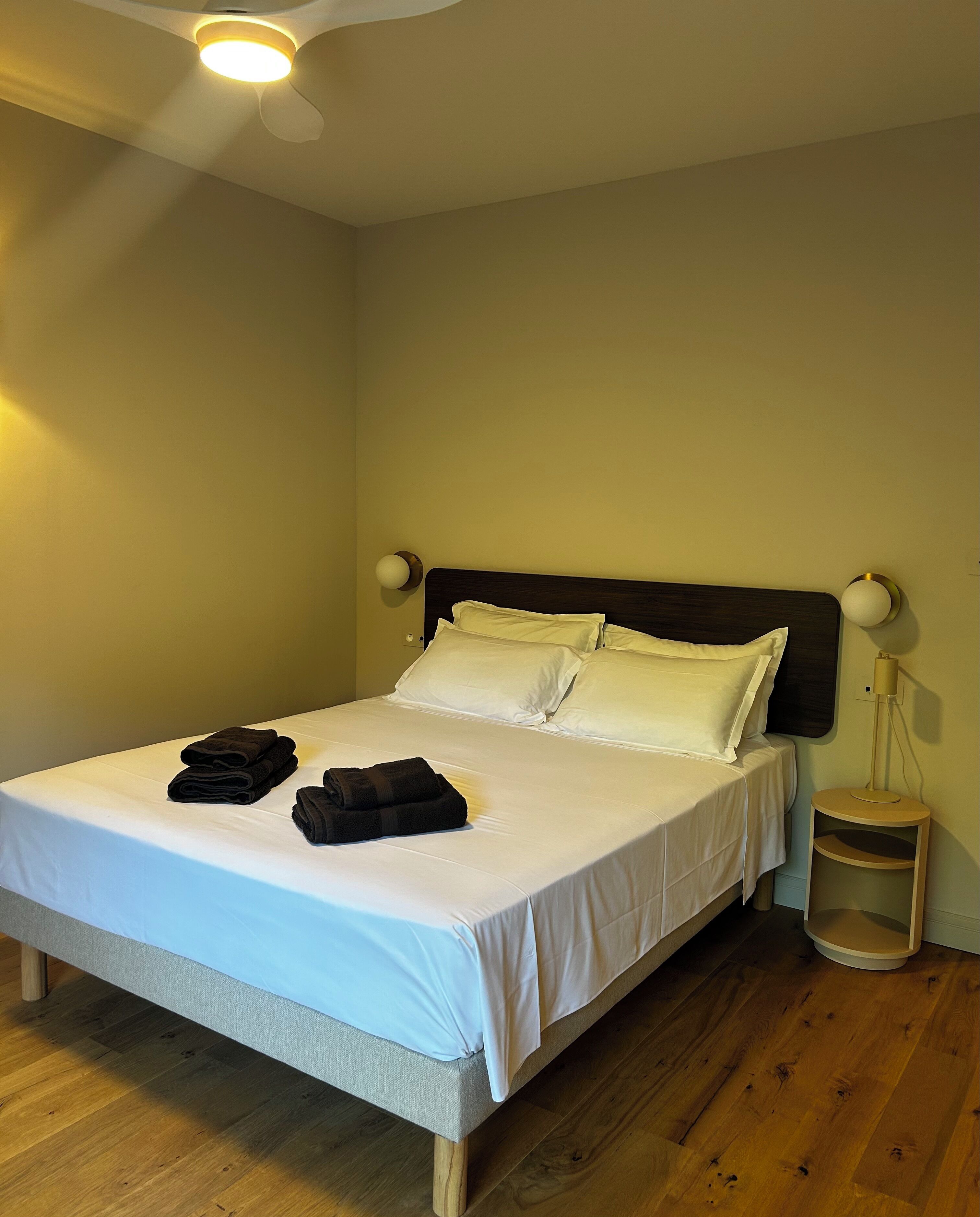 Una cama bien hecha con sábanas blancas y toallas dobladas, una lámpara de noche y suelo de madera.