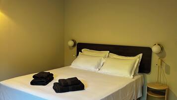 Una cama bien hecha con sábanas blancas y toallas dobladas, una lámpara de noche y suelo de madera.