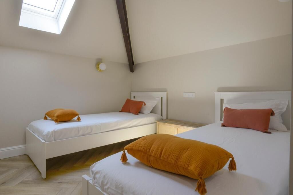 Un dormitorio con dos camas, cada una con ropa de cama blanca y almohadas naranjas. Hay un tragaluz sobre las camas y una mesita con una lámpara entre ellas.