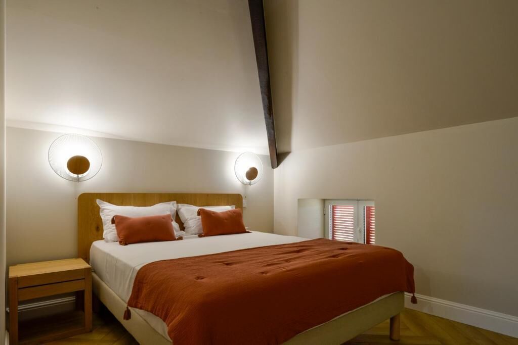 A bedroom with a bed, nightstand, wall-mounted lights, and a window with blinds.