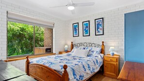 A bedroom with a wooden bed, two bedside tables, a window with a view of greenery, and a ceiling fan. - Coast and Country - 16 Riverview Drive, Carrickalinga (Carrickalinga)