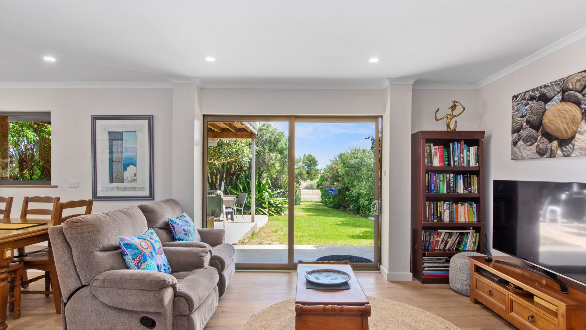 A modern living room with a sofa, coffee table, and bookshelf, a TV, and a sliding glass door leading to a garden.
