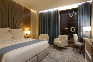 A hotel room with a large bed, two armchairs, a desk with a lamp, and a view of the interior through a glass door.