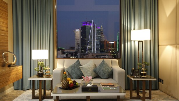 A hotel room with a cityscape view, a sofa, two bedside tables with lamps, a coffee table with fruits and a vase, and a mirror on the left wall. - Cantonal Hotel Riyadh (Riyadh)