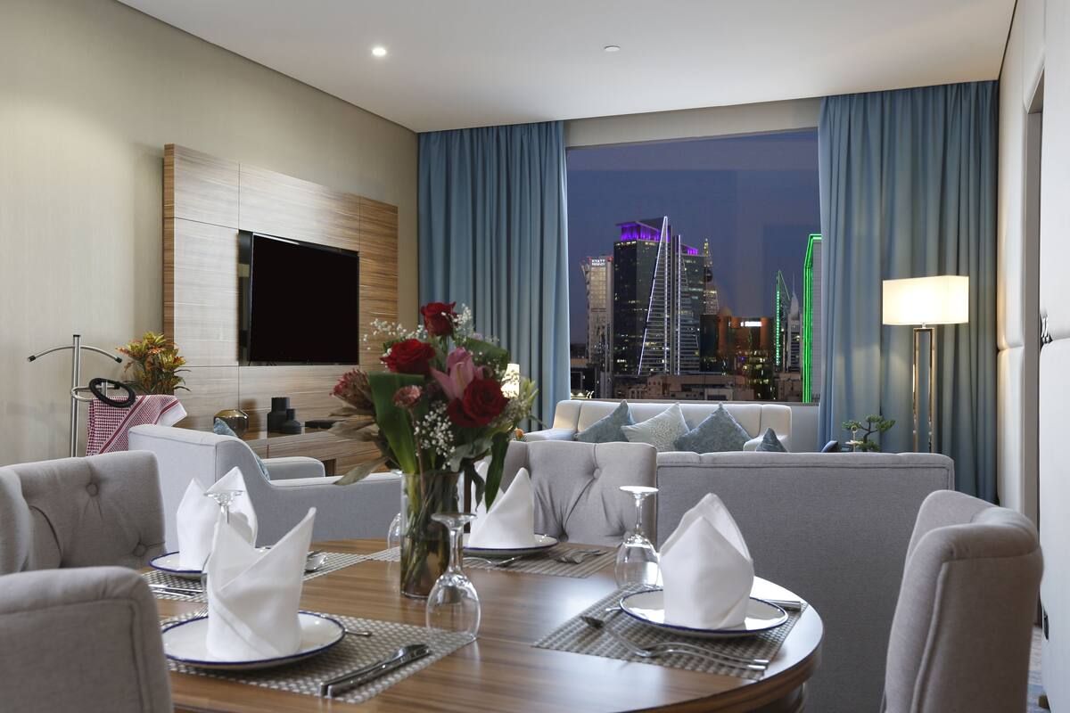 A hotel room with a dining area set for a meal, a sofa, a TV mounted on the wall, and a cityscape view through the window.