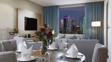 Ein Hotelzimmer mit Essbereich, gedecktem Tisch, einer Couch, einem an der Wand befestigten Fernseher und Blick auf die Stadt durch das Fenster.