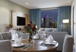 A hotel room with a dining area set for a meal, a sofa, a TV mounted on the wall, and a cityscape view through the window.