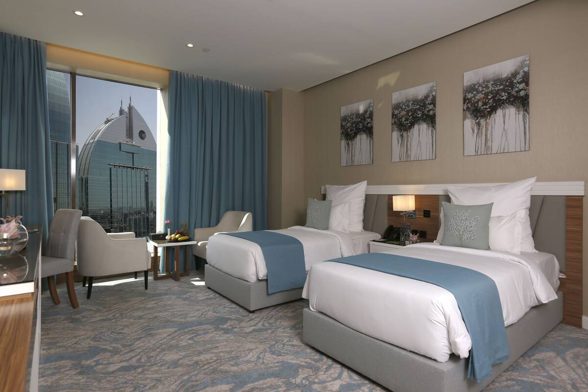 A hotel room with two beds, a desk, a chair, and a view of a building through large windows.