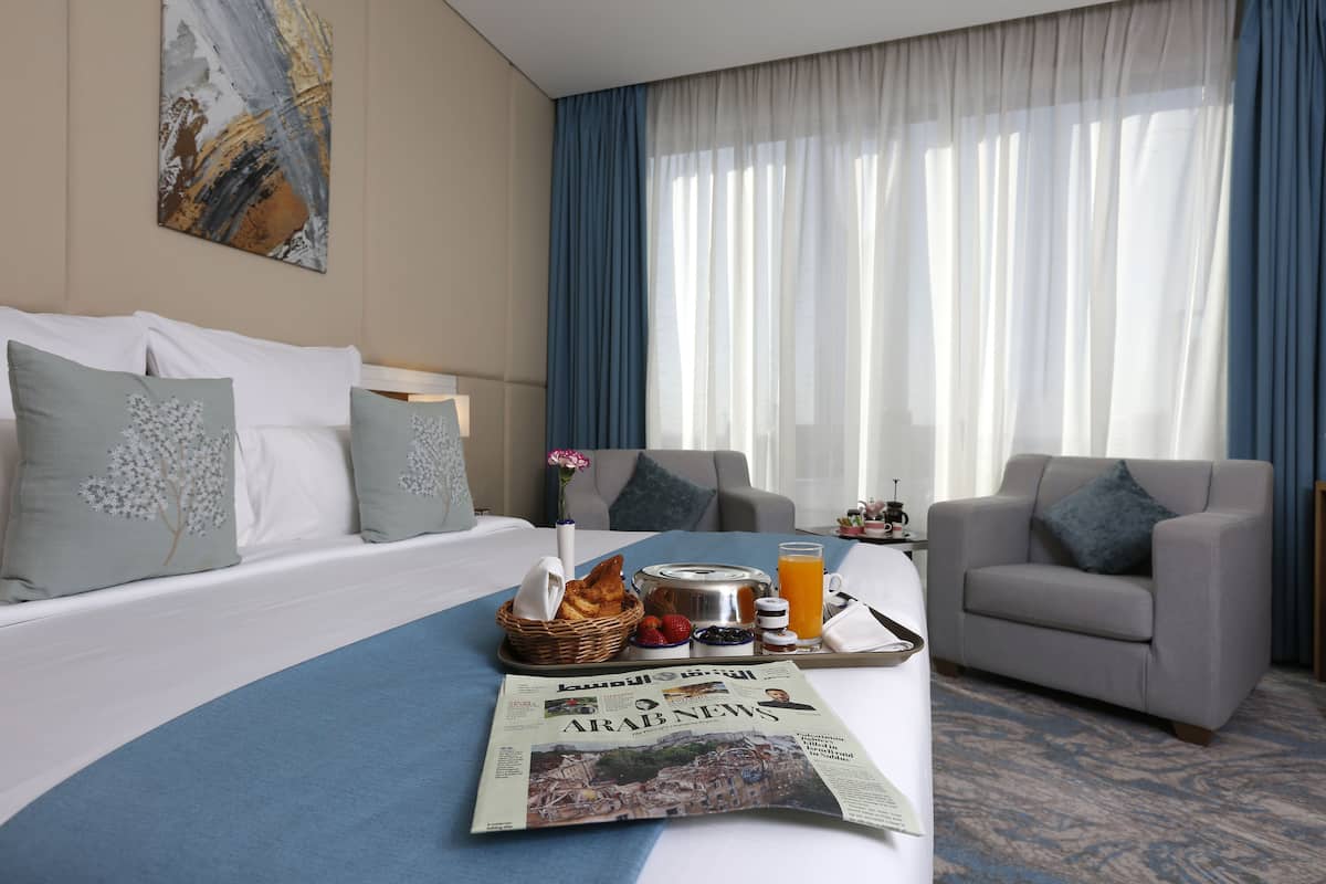 A hotel room with a bed, two armchairs, a breakfast tray with toast, fruit, and orange juice, and a newspaper.