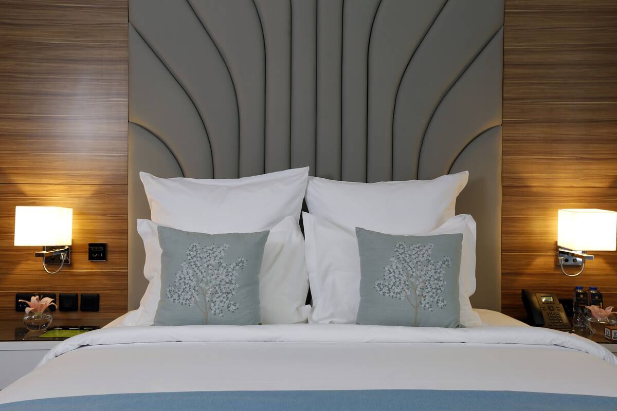 A neatly made bed with white and light blue pillows, a wooden headboard, bedside lamps, and a nightstand with a phone and a bottle.