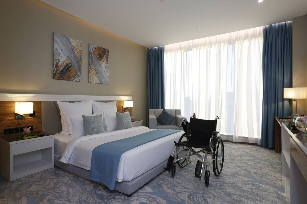 A hotel room with a large bed, a wheelchair, a nightstand, and a desk with a lamp.