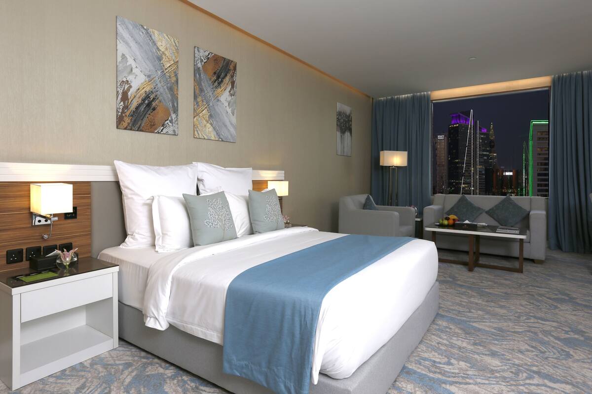 A hotel room with a bed, bedside tables, a desk, and a view of the cityscape at night.