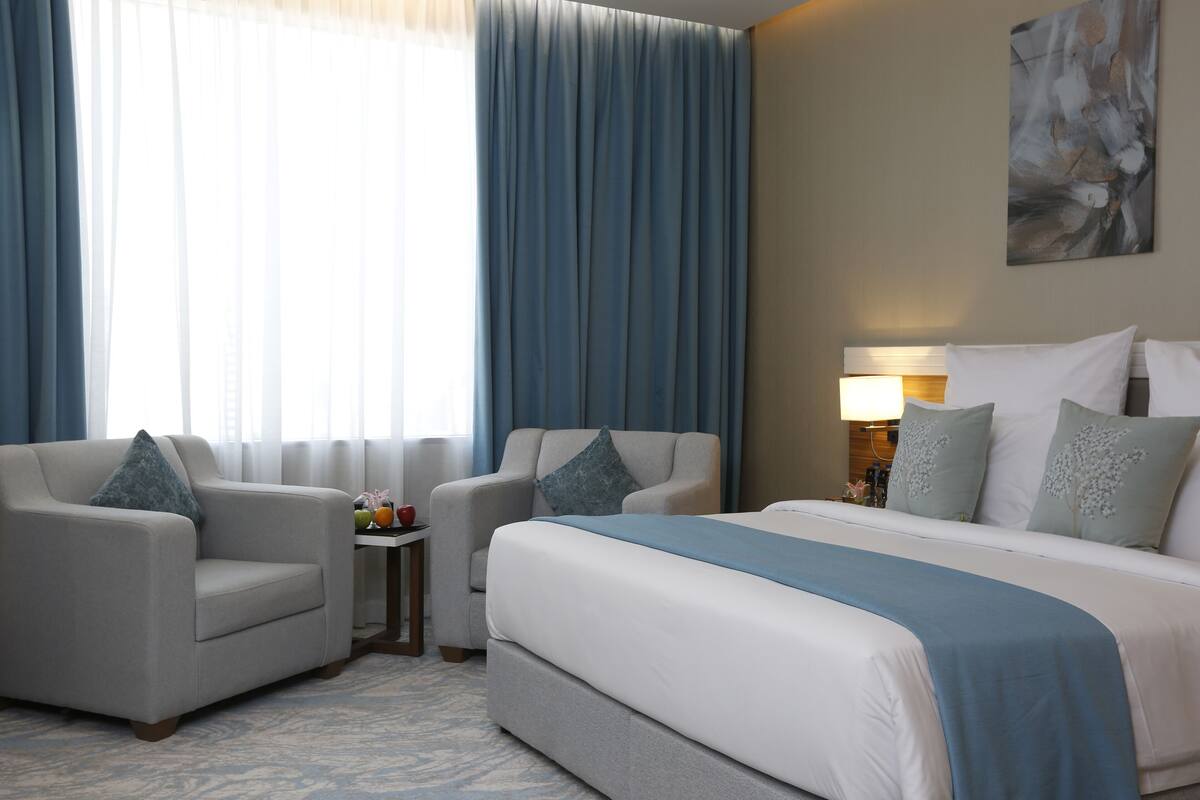 A hotel room with a bed, two armchairs, a small table with fruit, and a painting on the wall.