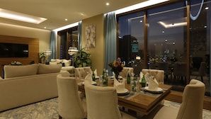 A modern dining area with a wooden table set for four, surrounded by upholstered chairs. A large window offers a view of the cityscape at night. - Cantonal Hotel Riyadh (Riyadh)