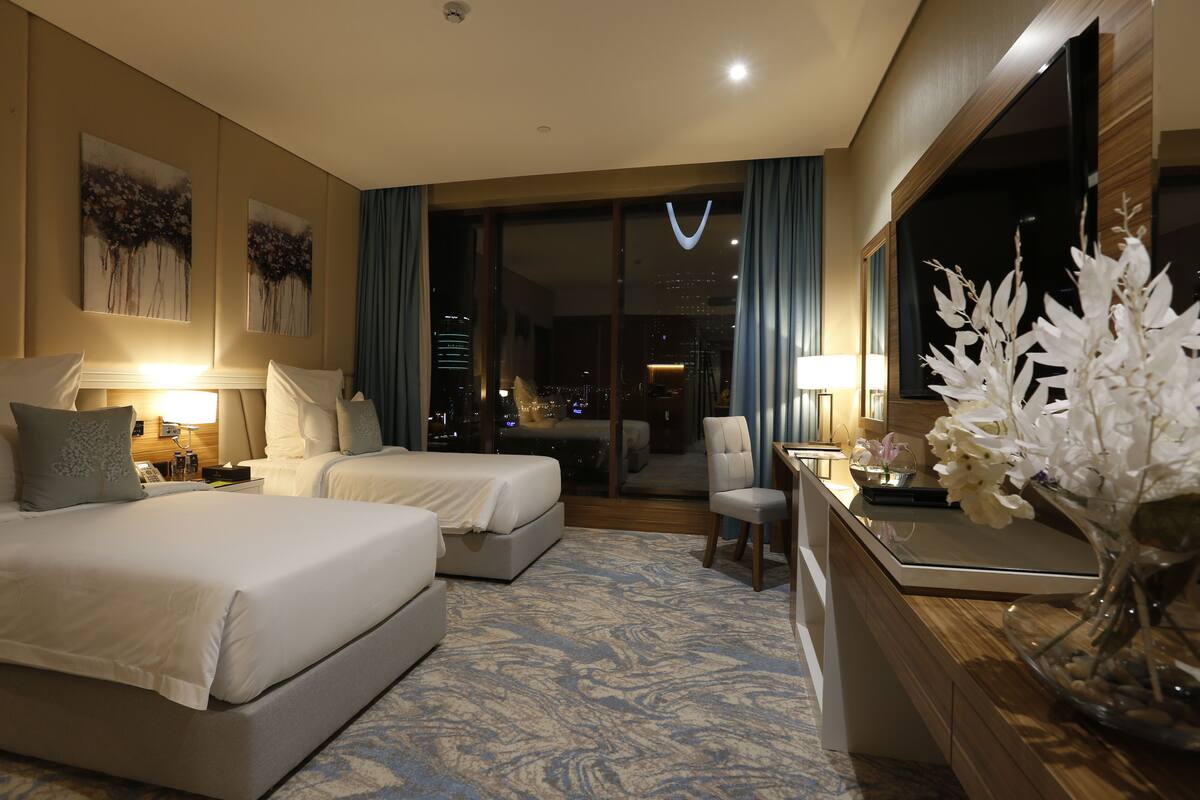 A hotel room with two beds, a desk, a chair, a large window with a city view, and a vase with flowers.