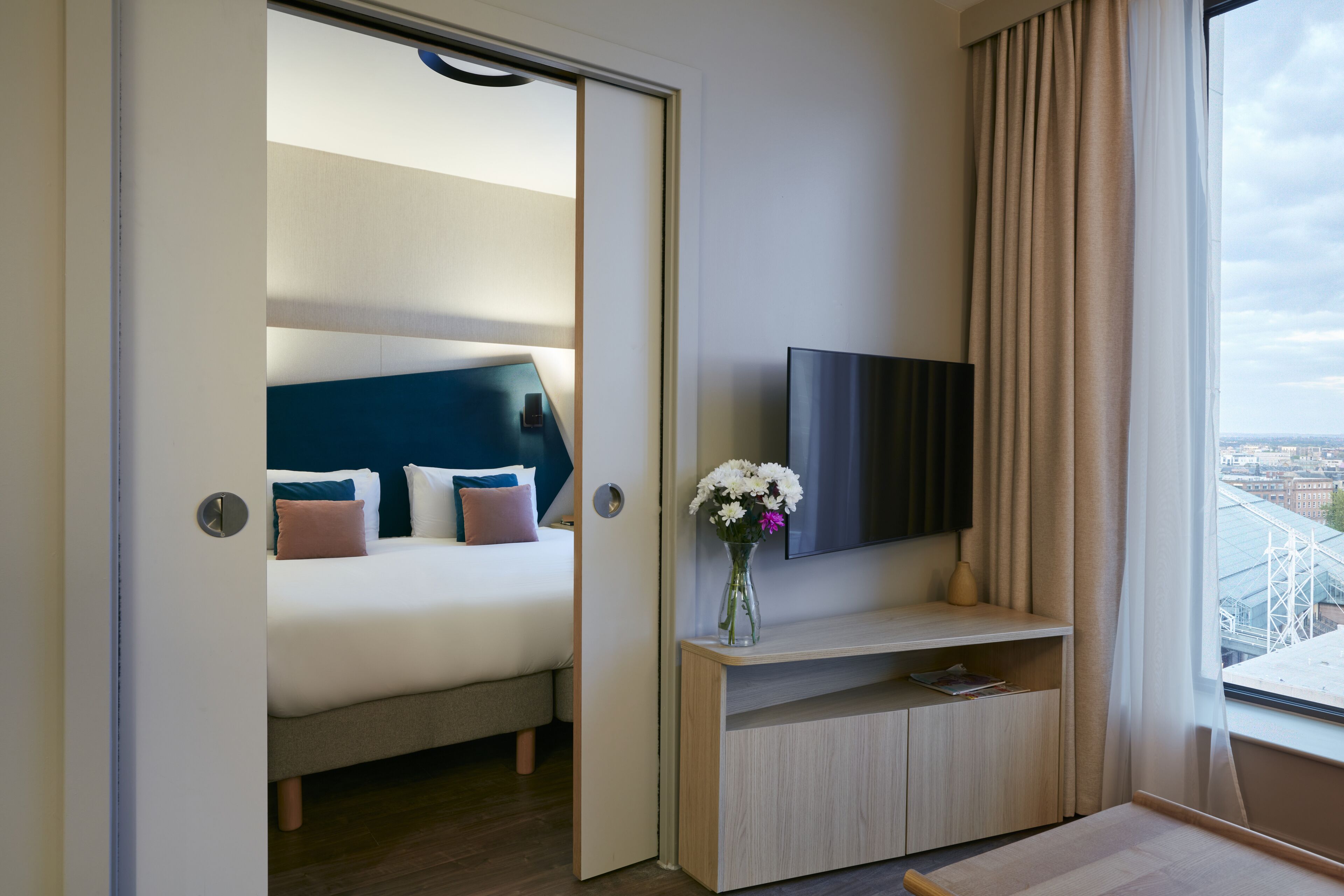 A hotel room with a bed, a TV, a vase with flowers, and a view of a bridge.