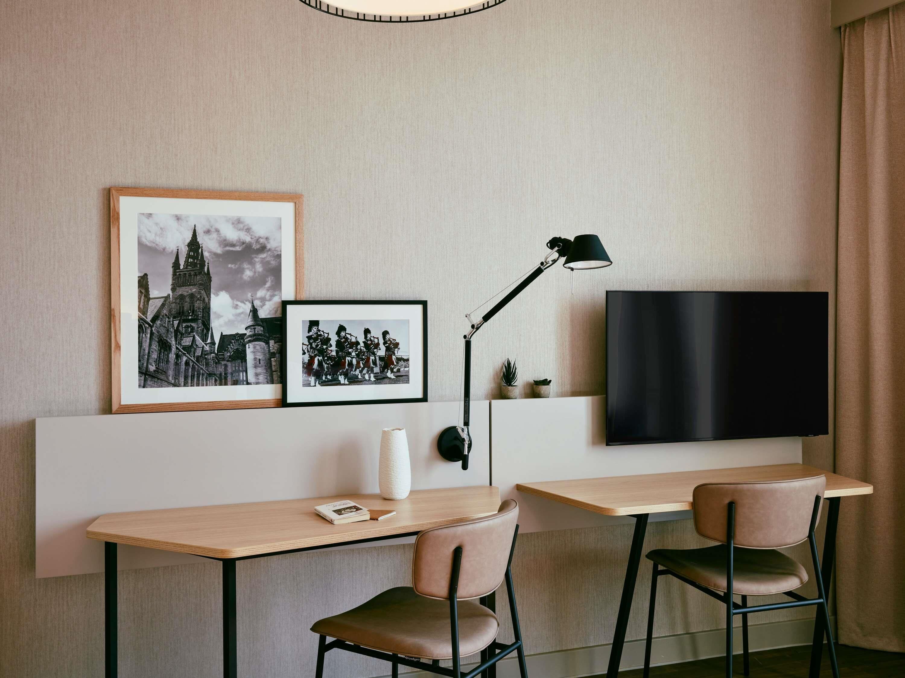 A modern hotel room with a flat-screen TV mounted on the wall, a desk with two chairs, and two framed pictures above the desk.