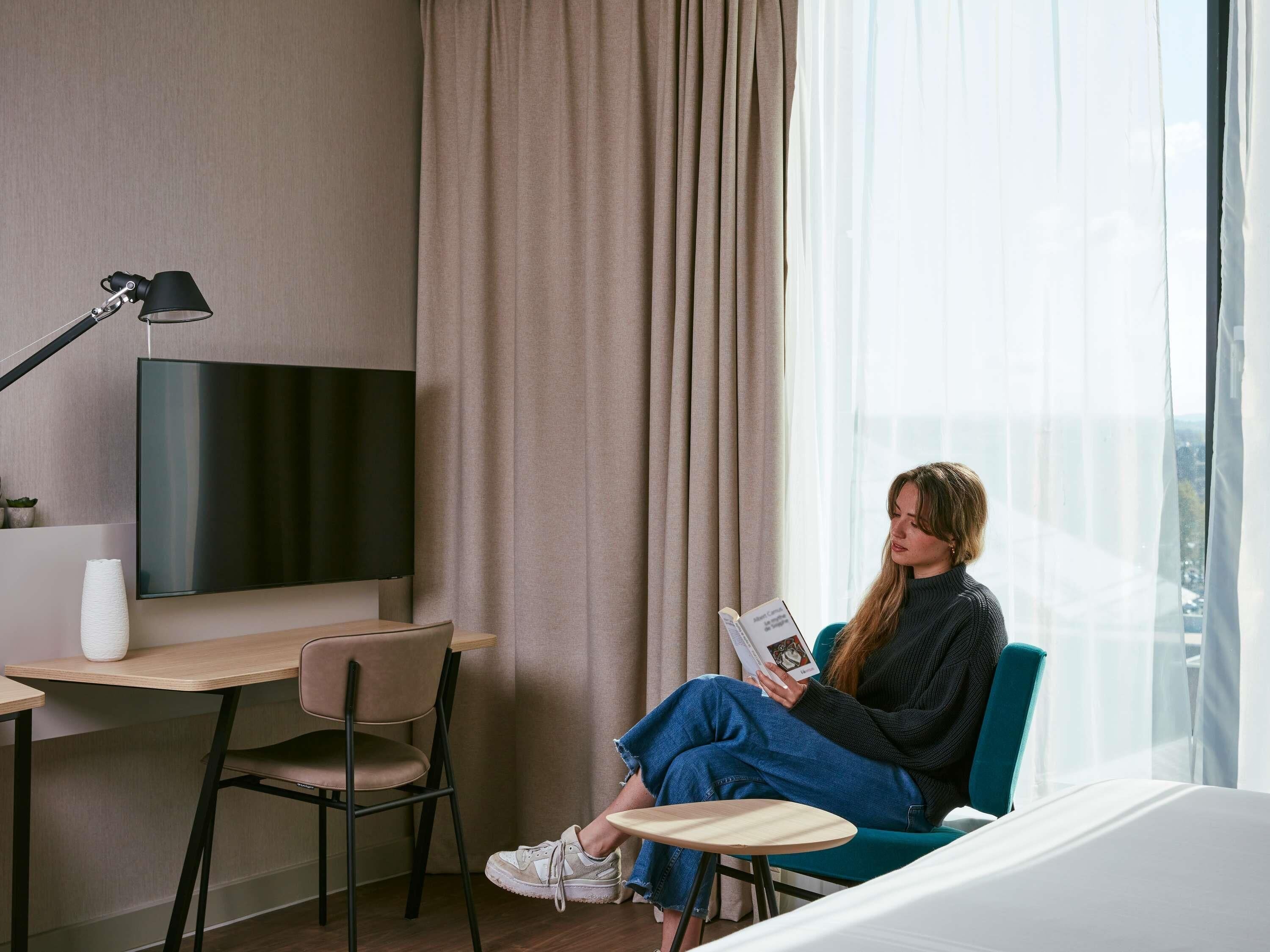 A modern hotel room with a flat-screen TV, a desk, a chair, and a window with curtains.