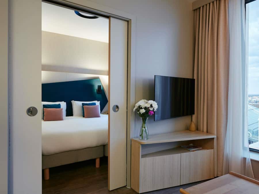 A hotel room with a bed, a TV, a vase with flowers, and a view of the city.