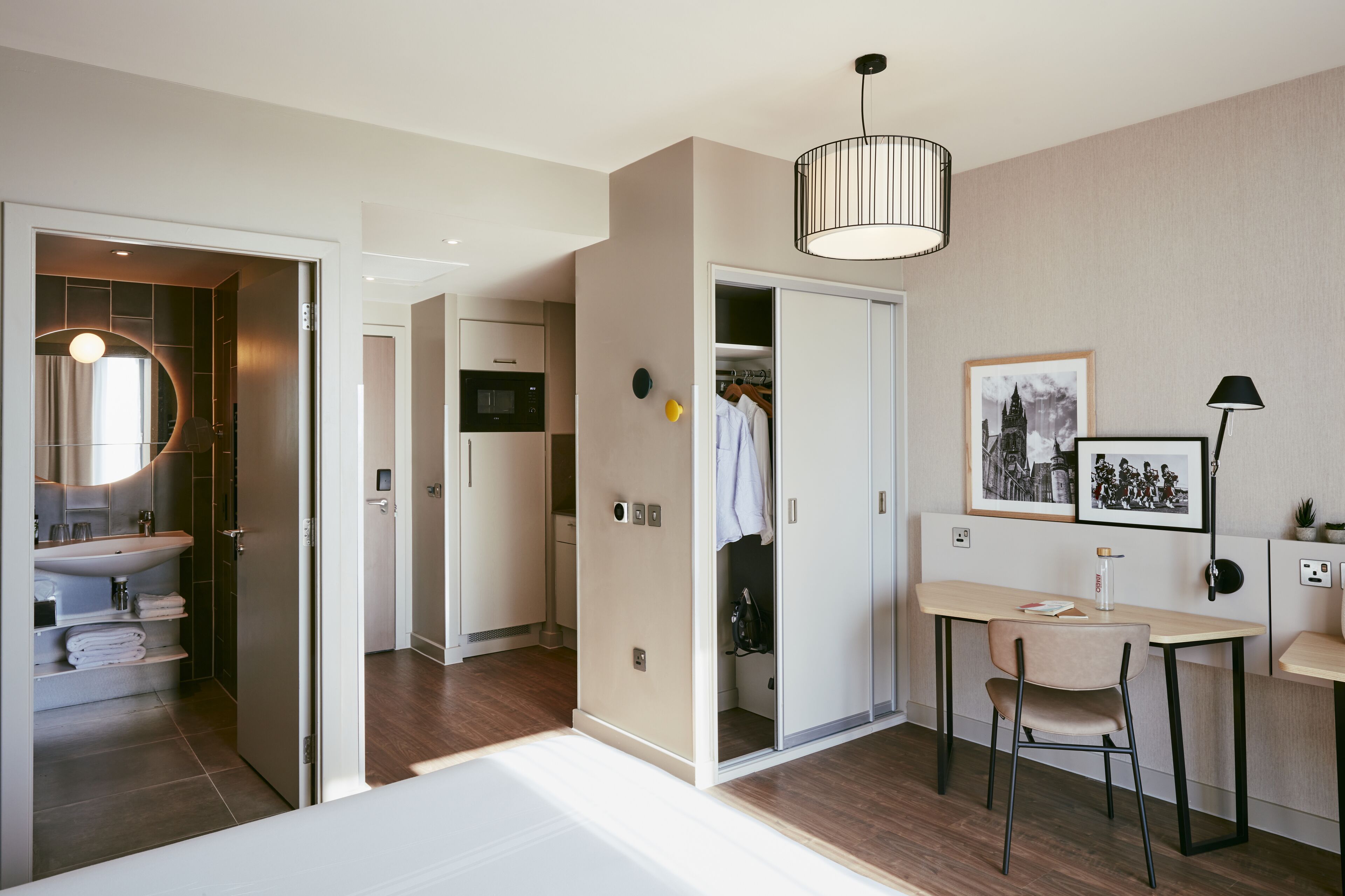 A modern hotel room with a bed, a desk with a chair, a wardrobe, a bathroom with a sink, and a mirror.