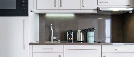 A modern kitchen with white cabinets, a stainless steel sink, a kettle, and a coffee maker.