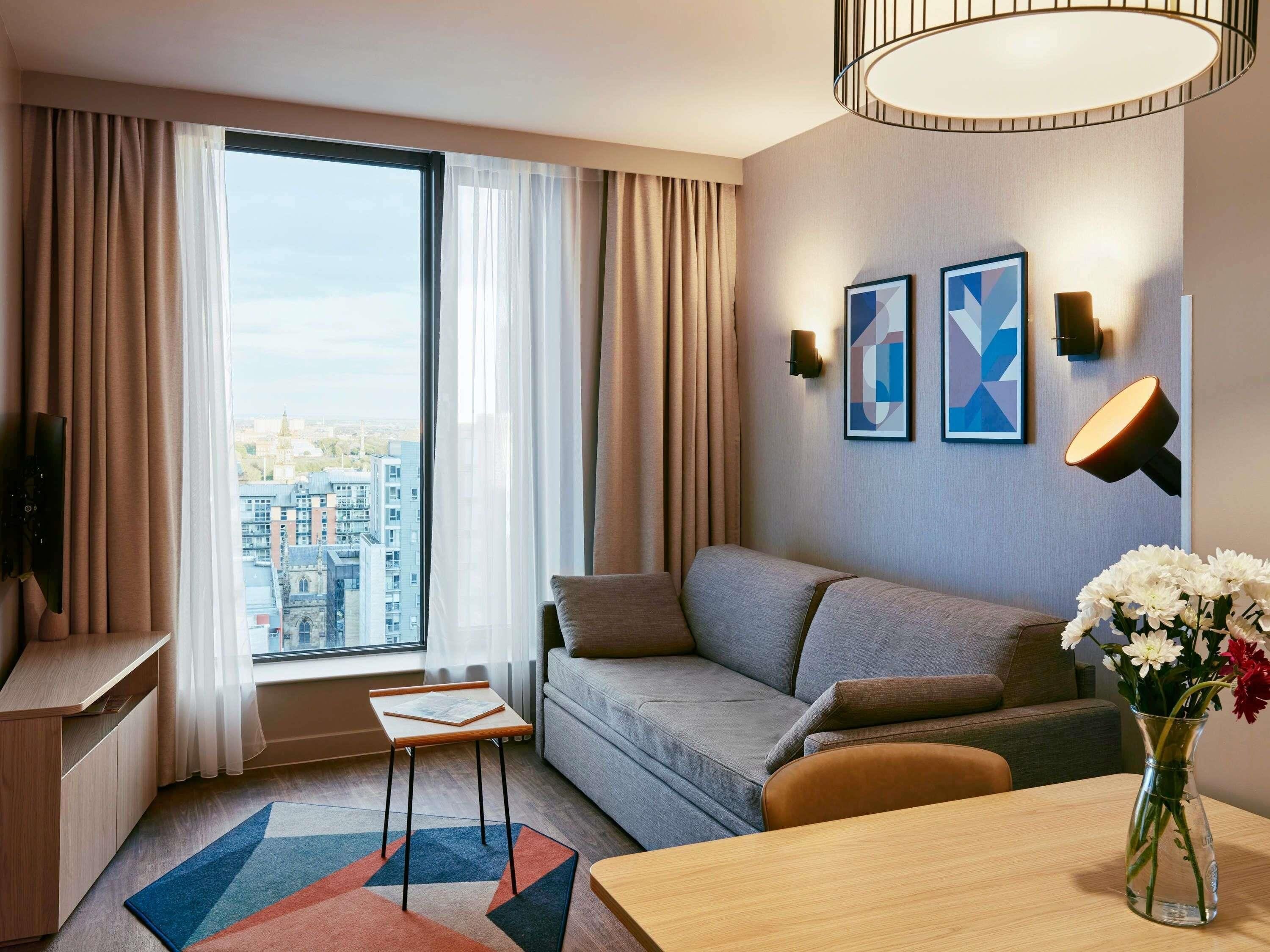 A modern hotel room with a sofa, a small table, a vase with flowers, and a city view.