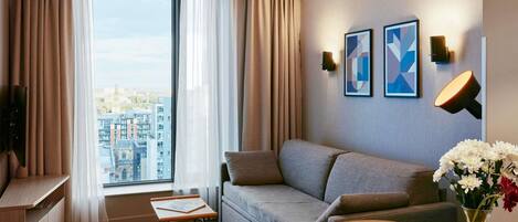 A modern hotel room with a sofa, a small table, a vase with flowers, and a city view.