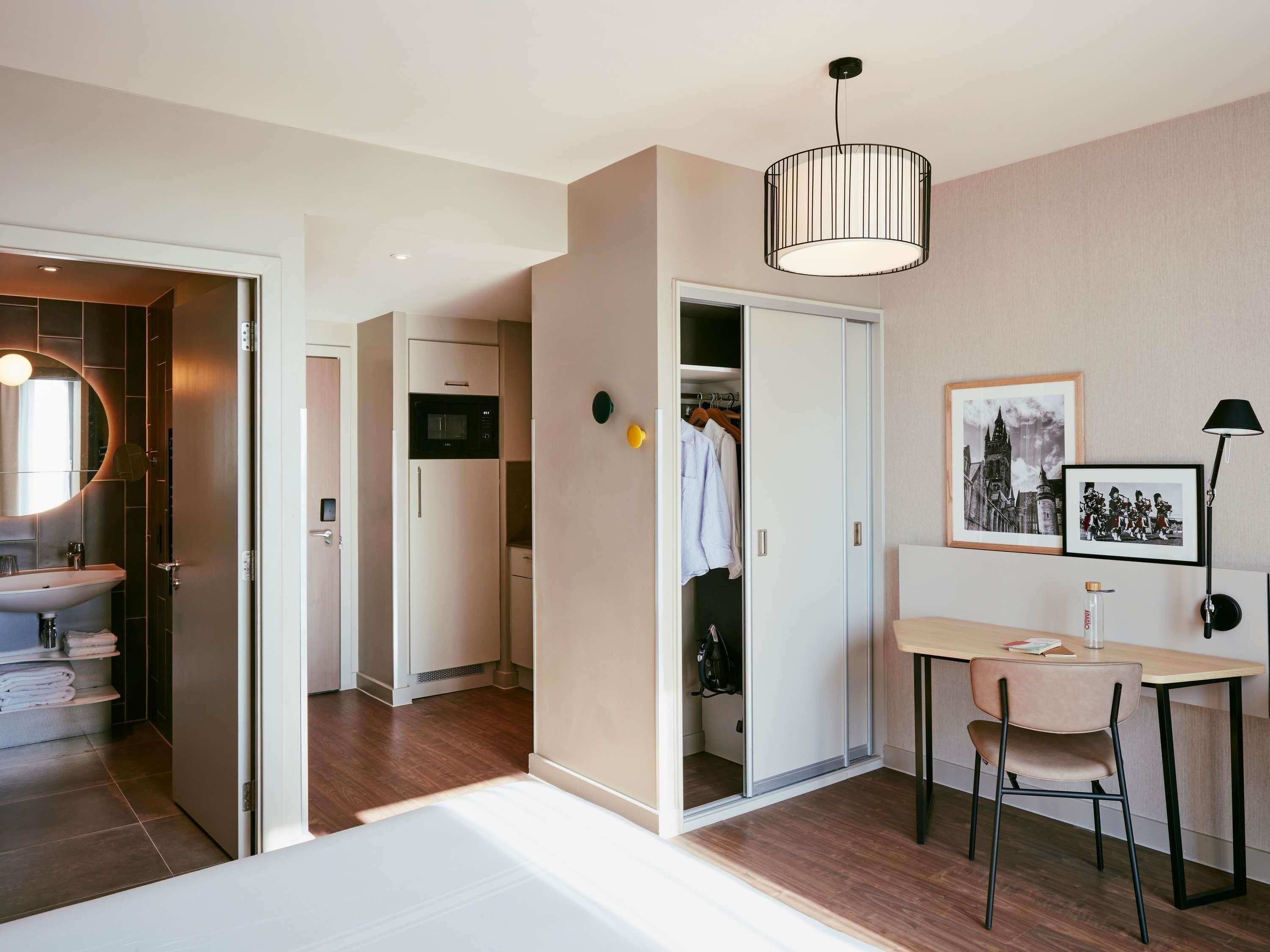 Ett hotellrum med en säng, ett skrivbord med en stol, en garderob, ett badrum med handfat och en mikrovågsugn.