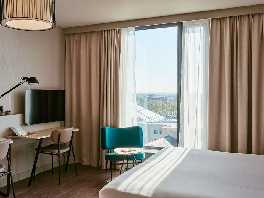 A hotel room with a bed, a desk, a chair, a TV, and a window with curtains.