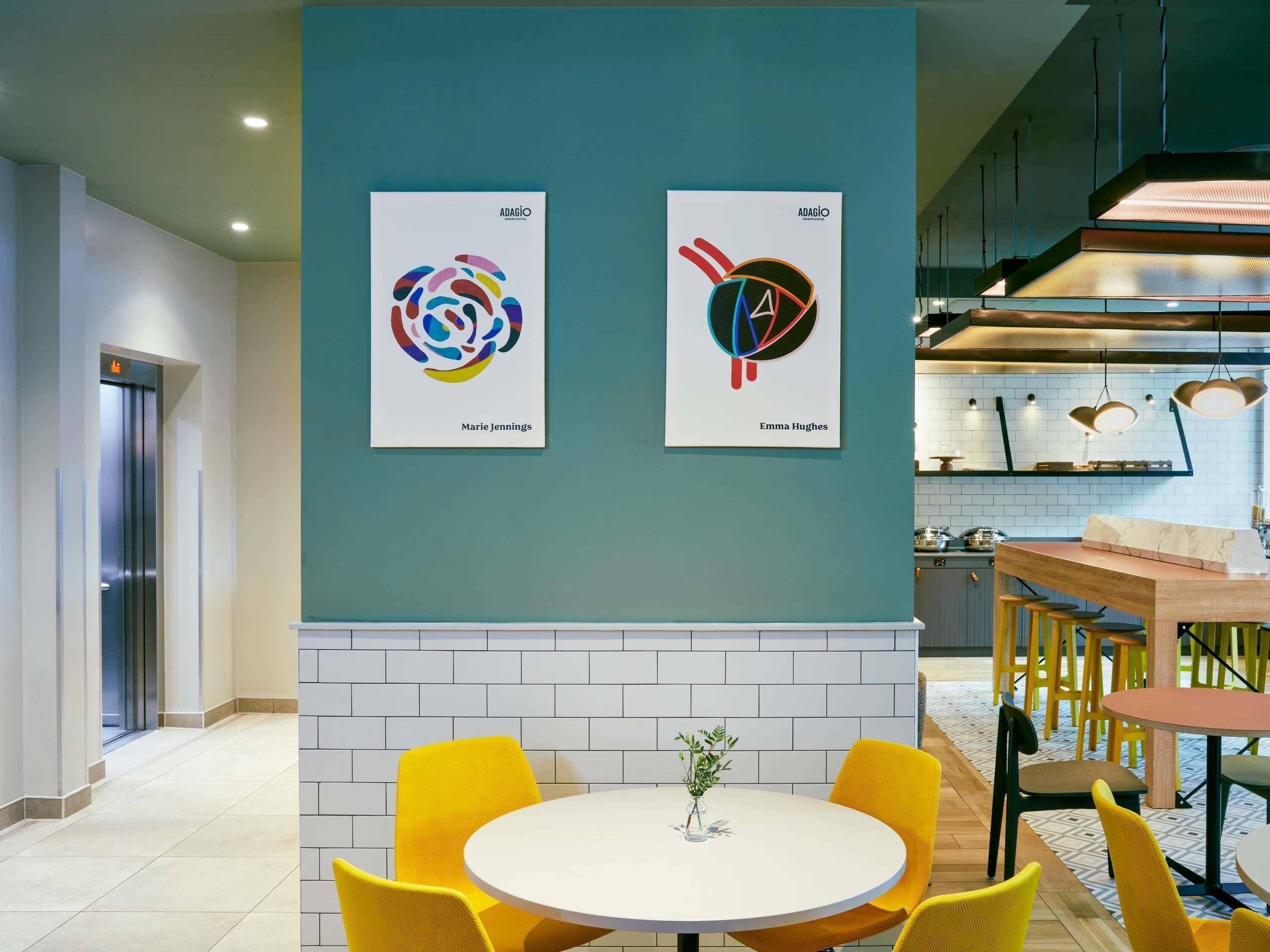 Un intérieur moderne agrémenté de deux œuvres d’art encadrées, d’un coin repas avec des chaises jaunes et d’une cuisine avec des luminaires suspendus.